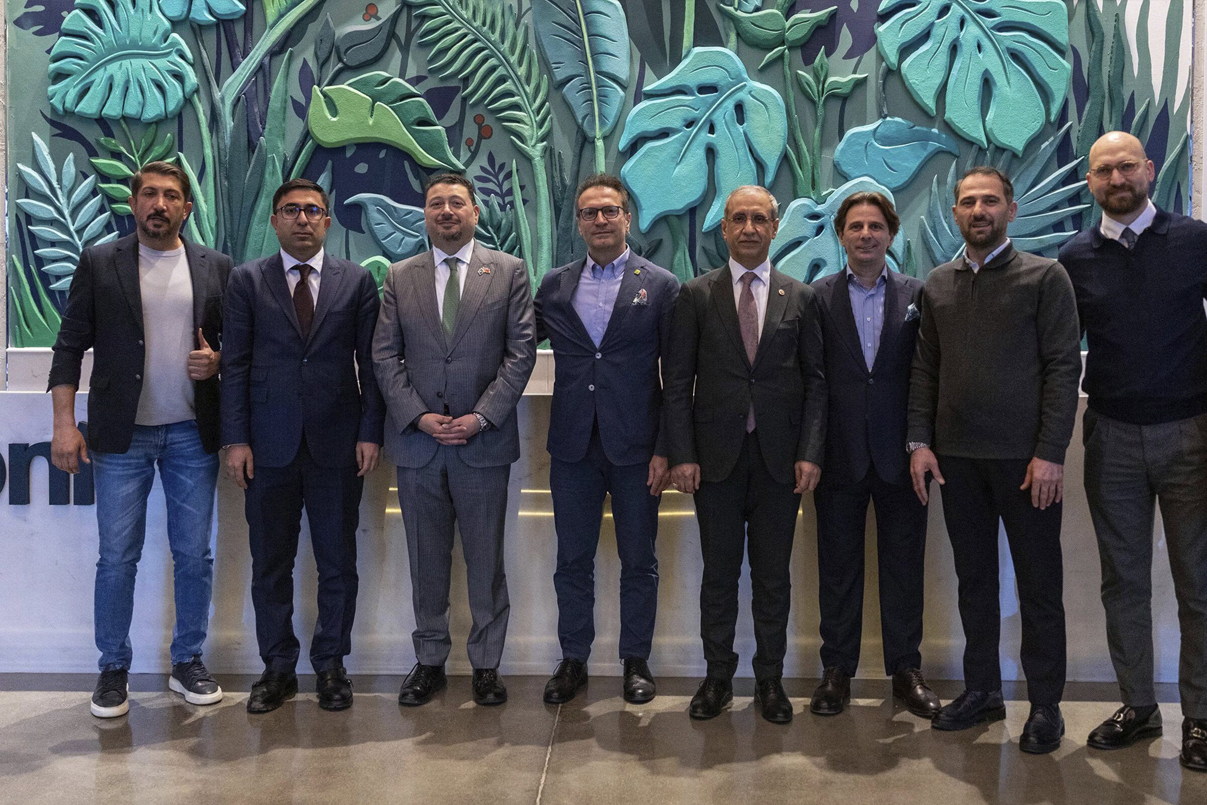 Freshly dressed professionals in suits standing in front of a vibrant, nature-inspired wall art at Saloni Furniture event, showcasing modern office decor and stylish interior design solutions for contemporary spaces.