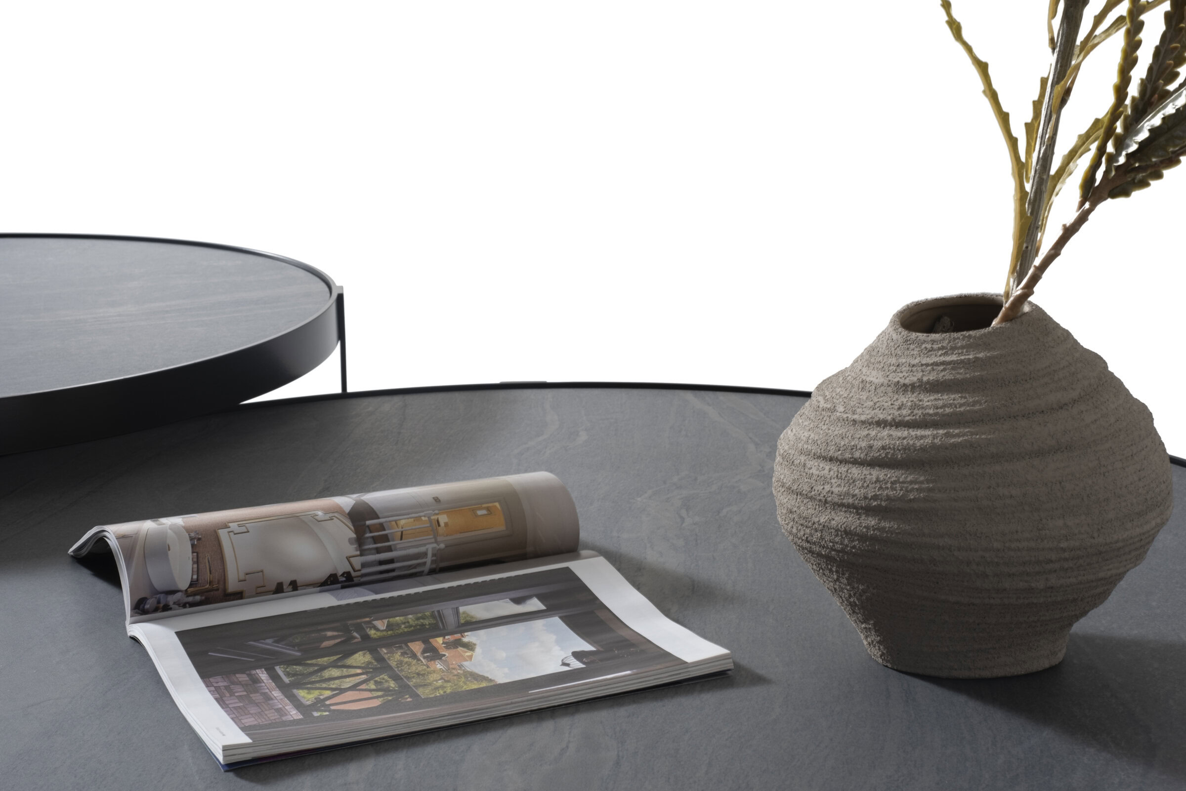 Modern textured beige ceramic vase with green foliage, placed on a sleek black round side table, accompanied by an open interior design magazine showcasing stylish home decor ideas.