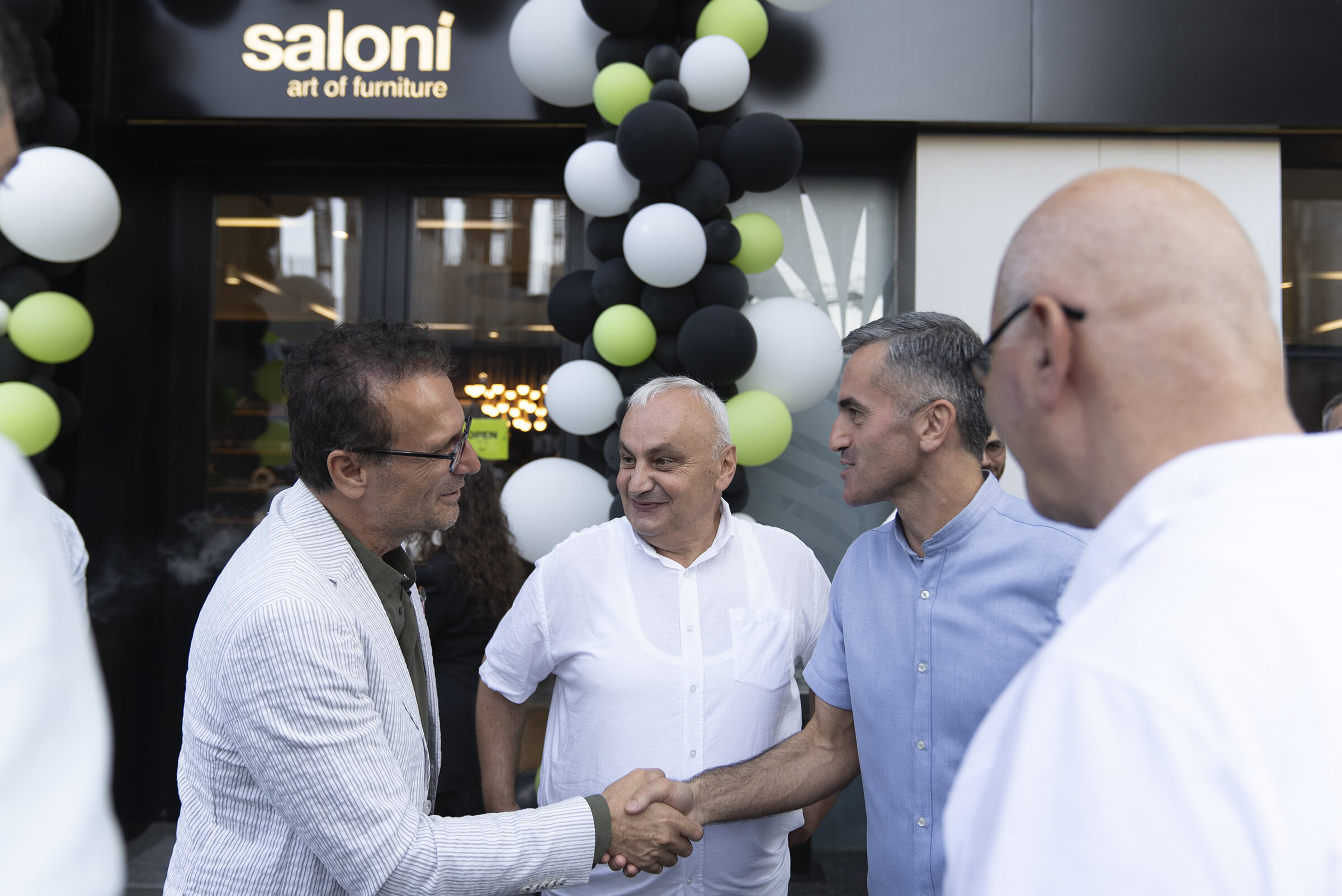 Modern furniture store grand opening with business professionals shaking hands outside Saloni Furniture, showcasing contemporary home and office furniture solutions, stylish decor, and inviting interior designs.