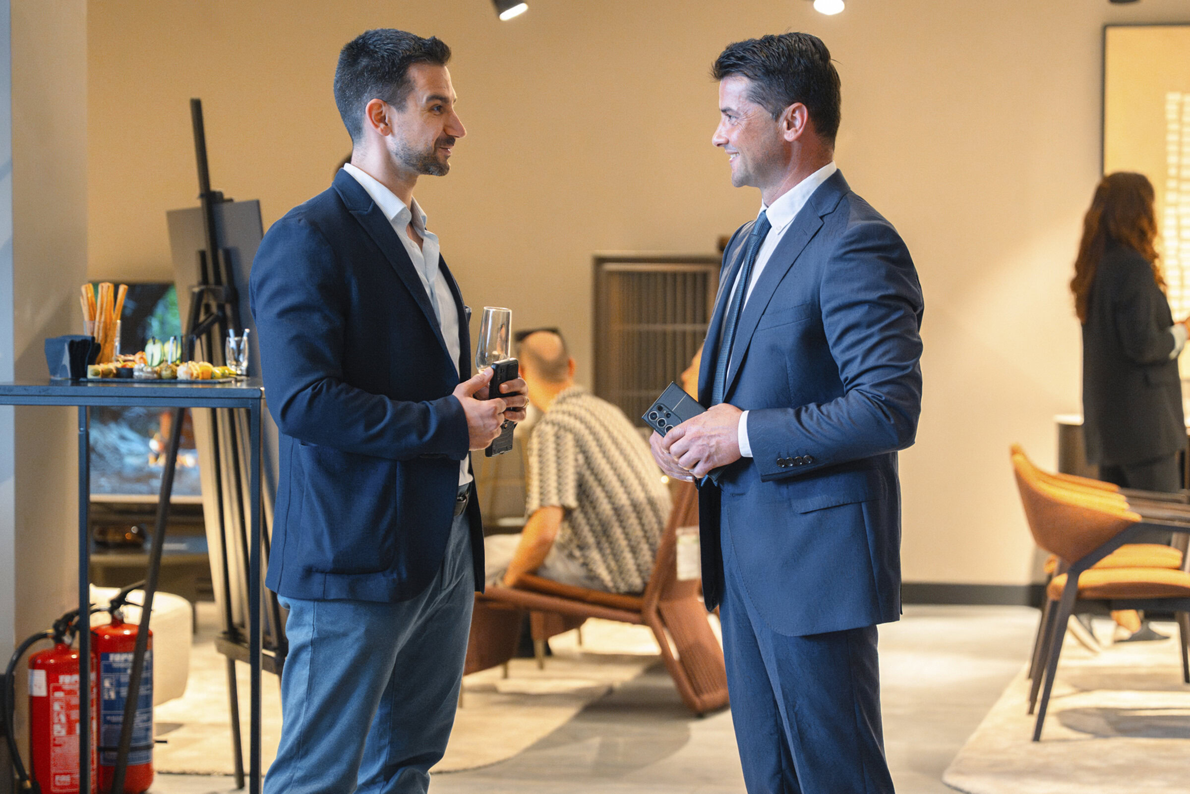 Modern office lounge with two businessmen in formal suits having a conversation, stylish seating area, contemporary furniture, and professional ambiance.