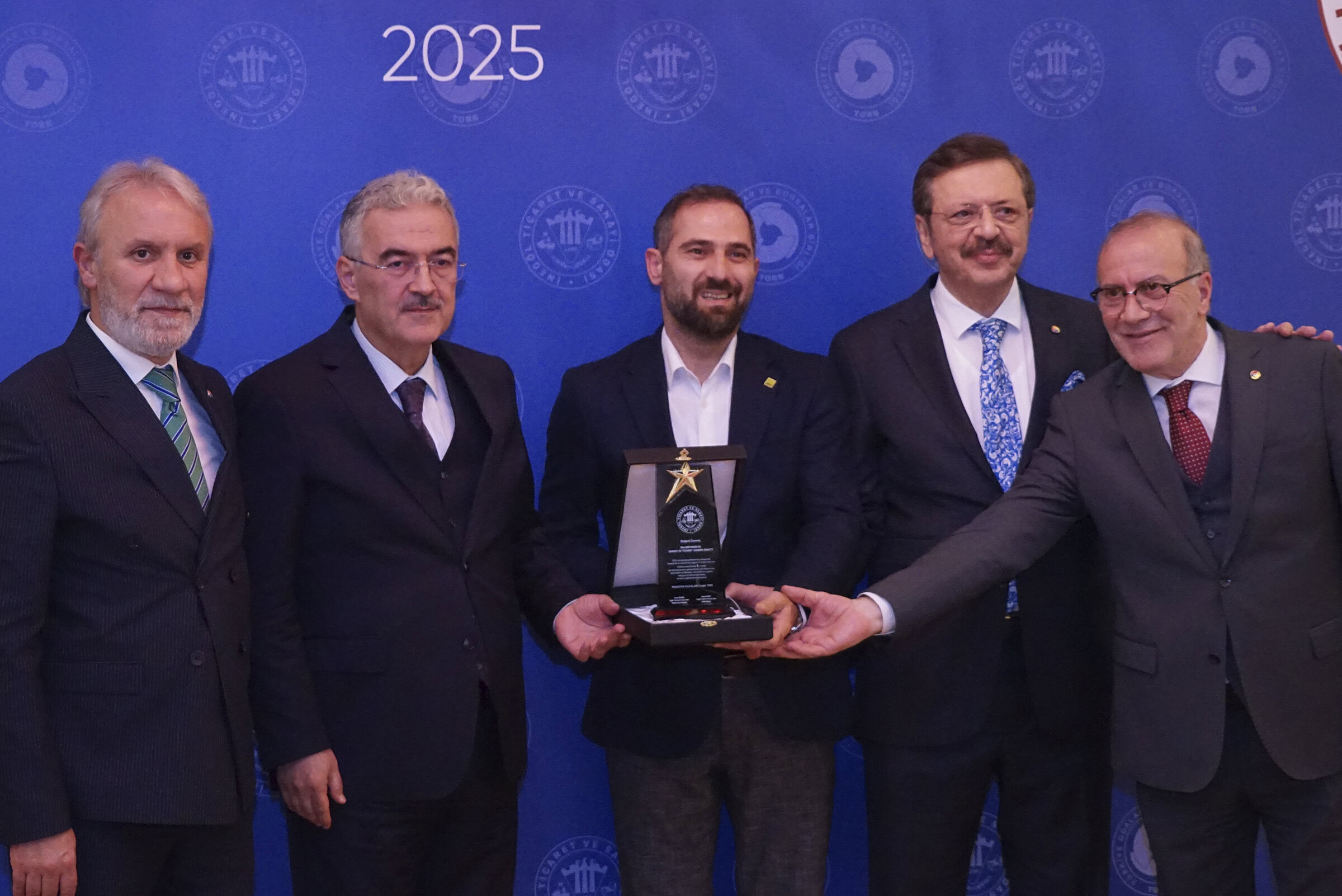 Handsome men in suits receiving an award at a formal event with a blue backdrop featuring logos and the year 2025, representing a professional and prestigious occasion.
